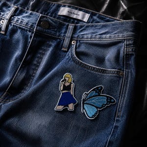 May include: Blue denim jeans featuring an Acne Studios label at the waistband. Two embroidered patches are attached to the front: one depicts a blonde figure in a black top and blue skirt holding a microphone, and the other is a blue butterfly.