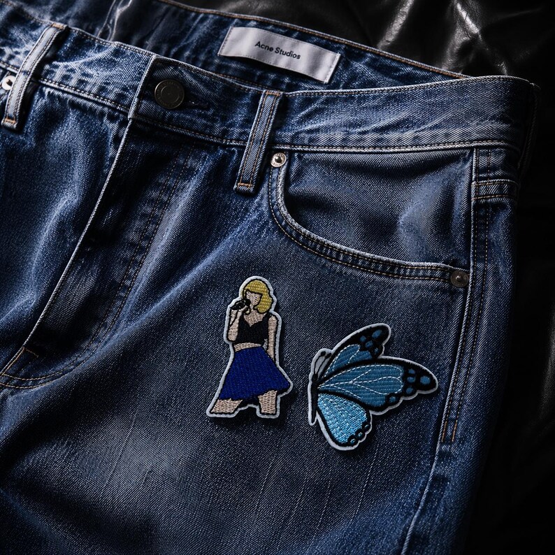May include: Blue denim jeans featuring an Acne Studios label at the waistband. Two embroidered patches are attached to the front: one depicts a blonde figure in a black top and blue skirt holding a microphone, and the other is a blue butterfly.