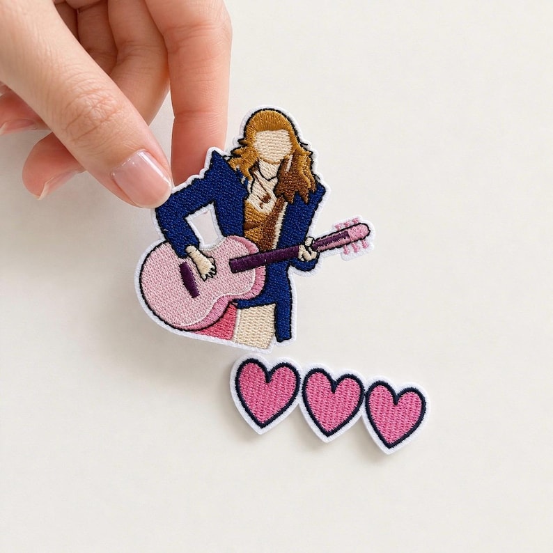 May include: An embroidered patch featuring a figure with brown hair wearing a dark blue jacket and playing a light pink acoustic guitar, accompanied by a separate smaller embroidered patch consisting of three connected pink hearts with dark outlines.