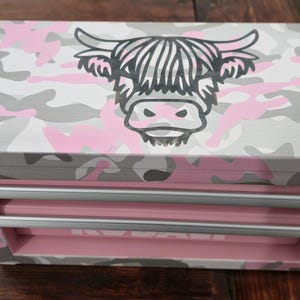 May include: A pink, gray, and white camouflage-patterned toolbox with three drawers. The top of the box features a black and white illustration of a Highland cow's head. The drawers and interior are pink.
