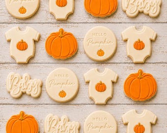 May include: A collection of sixteen decorated sugar cookies arranged in a grid. The cookies include round shapes with the text Hello Pumpkin, onesies with a small pumpkin design, orange pumpkins, and cookies with the script text Baby in white icing.