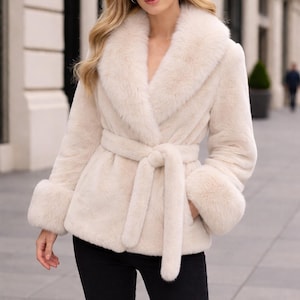 May include: A cream-colored faux fur jacket featuring a wide shawl collar, long sleeves with thick fur cuffs, and a matching tie belt at the waist. The jacket has a soft, plush texture and reaches the hip, providing a structured yet cozy appearance.