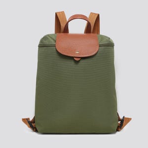 May include: An olive green backpack featuring a textured brown leather flap with a gold-tone snap closure. The bag includes a top handle, two adjustable brown shoulder straps, and a zippered main compartment, all constructed from durable fabric.