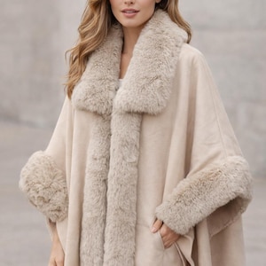 May include: A beige cape featuring a wide faux fur collar and matching faux fur trim along the front opening and sleeve cuffs. The garment has a smooth, solid-colored fabric body with integrated side pockets and a loose, open-front silhouette.