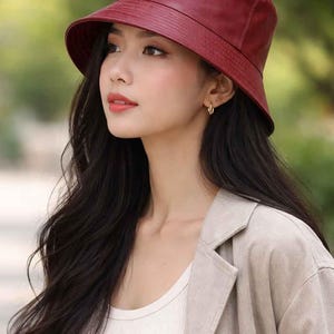 May include: A burgundy bucket hat with a structured brim. The hat is made of a smooth material and is worn by a person with long, dark hair. The person is also wearing a beige blazer and a white vest top.