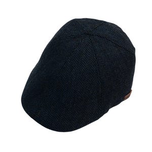 May include: A dark blue herringbone pattern newsboy cap. The cap has a rounded crown and a short, stiff brim. A small, brown leather accent is on the side. The cap is made of a textured fabric.