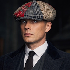 May include: A patterned flat cap with a red tartan section, and grey and tan check panels. The cap is worn with a dark grey suit, white collared shirt, and black tie. A gold tie bar is visible.
