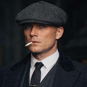 Op de afbeelding: Een man draagt een grijze tweed pet, een wit overhemd, een donkere stropdas en een donkergrijs pak. Een brandende sigaret zit in zijn mond. De foto is een portret met een donkere achtergrond.