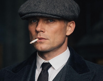 May include: A man wearing a gray tweed newsboy cap, a white collared shirt, a dark tie, and a dark gray suit. A lit cigarette is in his mouth. The image is a portrait with a dark background.