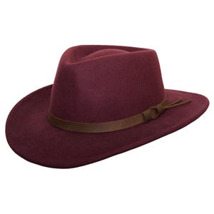 Puede incluir: Sombrero fedora burdeos con una banda de cuero marrón. El sombrero tiene un diseño clásico con ala ancha y corona pellizcada. La banda de cuero está atada con un nudo en el lateral. El sombrero es de material suave.