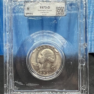May include: A 1973-D Washington Quarter coin encapsulated in a clear rectangular plastic slab. The label reads 1973-D Washington Quarter, Clashed Die, MS65. The coin features the profile of George Washington, the word LIBERTY, and IN GOD WE TRUST.