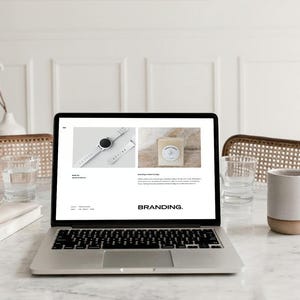 May include: A laptop displaying a website with images of a watch and soap. The word "BRANDING" is displayed on the screen. A mug, glass of water, and a book are on the marble table. A vase with flowers is in the background.