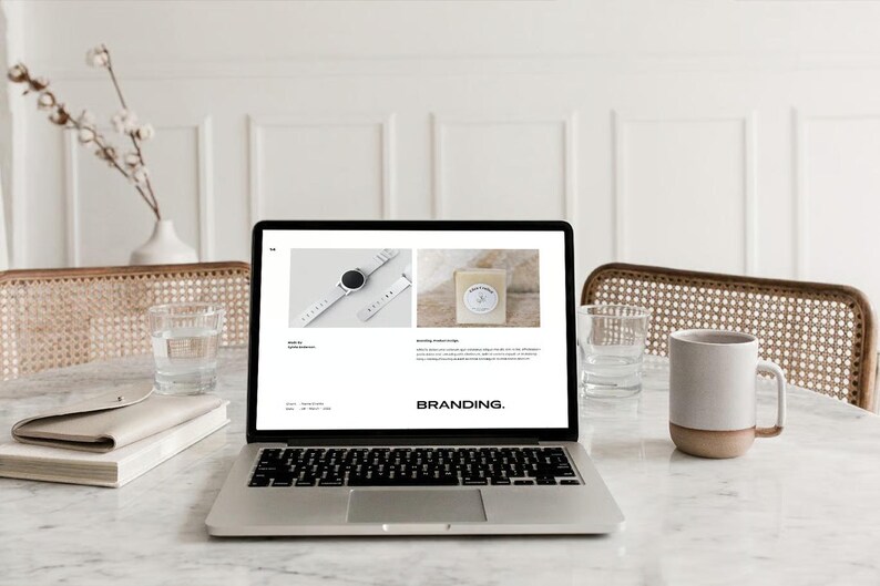 May include: A laptop displaying a website with images of a watch and soap. The word "BRANDING" is displayed on the screen. A mug, glass of water, and a book are on the marble table. A vase with flowers is in the background.