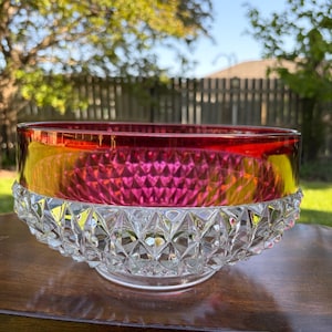 May include: A clear glass bowl with a ruby red and yellow rim. The bowl has a diamond-cut pattern on the lower half and sits on a clear base. The bowl is on a wooden table, with a green lawn and trees in the background.
