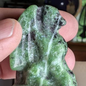 May include: A green and black carved stone figurine, possibly a small sculpture or decorative item. The stone has a mottled appearance with shades of green and black. The figurine is held between fingers, showcasing its size and detail.