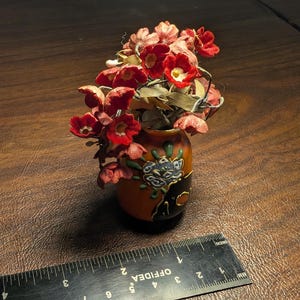 May include: A small, decorative vase with a floral arrangement. The vase is orange and black with a painted floral design. The flowers are red with white centers. A ruler is in the foreground.