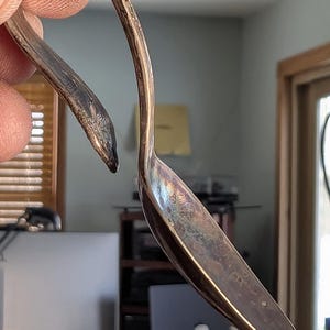 May include: A vintage silver spoon with a curved handle and a tarnished bowl. The spoon is held up, showcasing its unique shape and aged appearance. The spoon's design is reminiscent of a swan's neck.