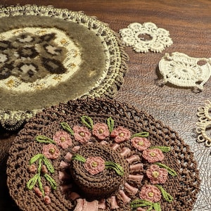 May include: A brown crocheted hat with pink floral embroidery and a pink ribbon. The hat features a circular design with a central floral motif and is surrounded by additional floral accents and green leaves. Decorative lace pieces are also visible.