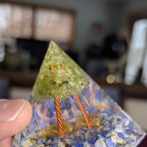 May include: A clear pyramid-shaped crystal with a green stone and gold glitter inside. Two copper wires are visible inside the pyramid.