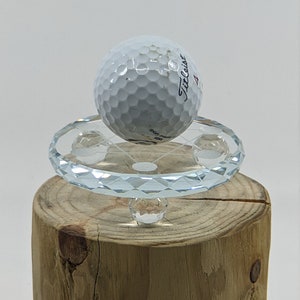 May include: A white golf ball with the text "Titleist" on it is balanced on a clear crystal pedestal with a wooden base.
