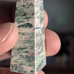May include: A gemstone obelisk with a faceted top. The stone features a mix of green, white, and pink hues, with a marbled appearance. The obelisk is held in a person's hand.