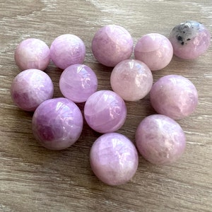 May include: A collection of twelve amethyst spheres, ranging in size, with a light purple hue and a smooth, polished finish. The spheres are arranged on a light brown wooden surface.