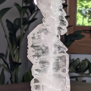 May include: A tall, clear, multi-faceted quartz crystal cluster standing vertically. The crystal features sharp, jagged edges and a translucent, glass-like appearance. It is securely mounted into a smooth, rounded, light gray stone base.