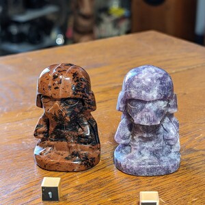 May include: Two small carved stone figurines of a character wearing a helmet. The figurine on the left is brown with black markings. The figurine on the right is purple. Both figurines are numbered 1 and 2.