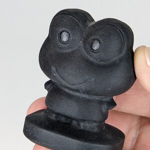May include: A black ceramic figurine of a frog with a wide smile and large eyes. The frog is standing on a small base.