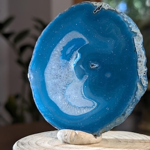 May include: A circular blue agate slice featuring a crescent-shaped white crystalline center. The stone has a polished, translucent surface with deep blue tones and a natural, rough, textured outer edge, displayed upright on a small stone base.