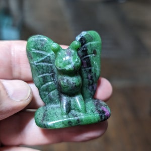 May include: A green and black carved stone figurine of a butterfly with wings outstretched. The figurine is made of a green stone with black and pink streaks.