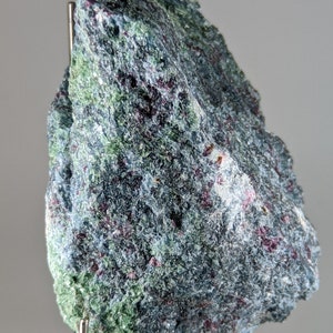 May include: A dark green and purple rock with a rough, textured surface. The rock is mounted on a silver stand.