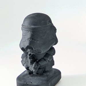 May include: A black 3D printed figurine of a Star Wars Stormtrooper. The figurine is in a crouching pose with its arms crossed in front of it. The figurine is on a black base.