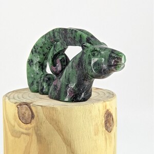 May include: A green and black carved gemstone sculpture of a goat's head with curled horns. The sculpture is sitting on a wooden base.