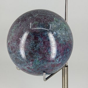 May include: A polished, dark blue and purple sphere with a speckled pattern, displayed on a metal stand with a spring.