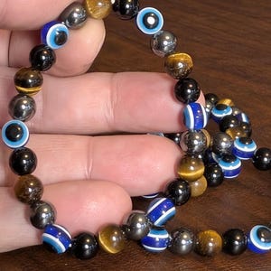 May include: A beaded bracelet featuring a variety of beads in black, gold, silver, and blue. The bracelet includes beads with an eye design, tiger's eye beads, and metallic beads. The bracelet is held in a hand.