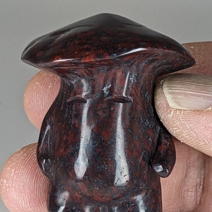 May include: A hand-carved figurine of a person with a large, rounded head and a wide, flat brimmed hat. The figurine is made of a dark red and black stone with a speckled pattern.