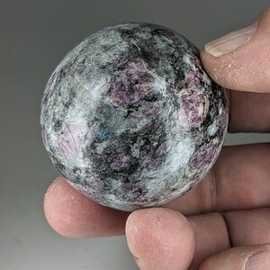 May include: A smooth, round, grey and black sphere with pink and purple flecks. The sphere is made of polished stone.