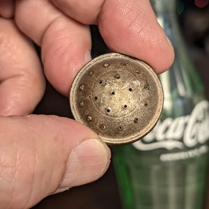May include: A small, round, antique metal object with numerous small holes, held between fingers. The object has a slightly domed shape and a tarnished, aged appearance. The background includes a green glass bottle.