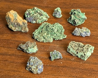 May include: A collection of raw azurite and malachite mineral specimens. The stones vary in size and shape, displaying a mix of deep blue and vibrant green hues. The minerals are arranged on a brown surface.
