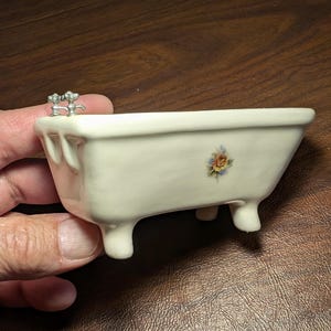 May include: A small, off-white ceramic bathtub with four rounded feet and a silver faucet. A floral design with orange, yellow, and blue flowers decorates the side. The miniature tub is held by a hand.