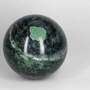 May include: A smooth, dark green sphere with a lighter green patch on the surface. The sphere is made of polished stone.