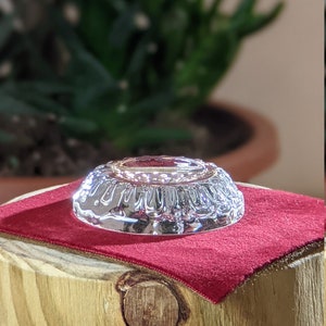 May include: A silver-colored, round, decorative dish with a raised, textured rim. The dish is sitting on a red velvet surface.