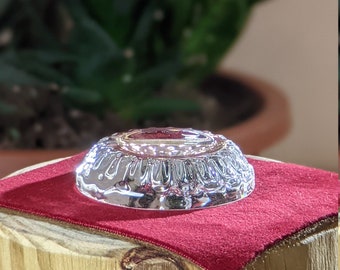 May include: A silver-colored, round, decorative dish with a raised, textured rim. The dish is sitting on a red velvet surface.