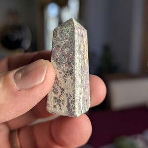 May include: A small, pointed, green and white crystal with pink and purple flecks. The crystal is in the shape of a pyramid.