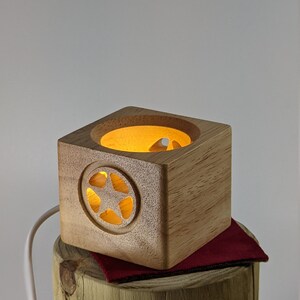 May include: A small wooden cube with a star-shaped cutout and a warm light glowing from inside. The cube is sitting on a wooden stump with a red fabric underneath.