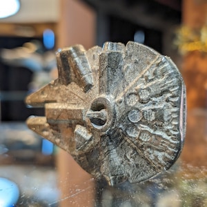 May include: A silver-colored metal model of the Millennium Falcon spaceship from Star Wars. The model is detailed with textured surfaces and a small opening for the cockpit.