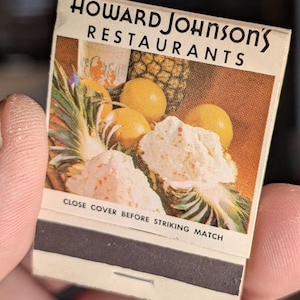 May include: Vintage matchbook cover for Howard Johnson's Restaurants. The cover features a colorful illustration of a pineapple, lemons, and a dessert. Text reads "Howard Johnson's Restaurants" and "Close Cover Before Striking Match."