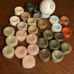 May include: A collection of small, polished stone golf ball markers in various colors, including pink, green, and brown. A white golf ball is also present. The markers are arranged on a dark brown surface.