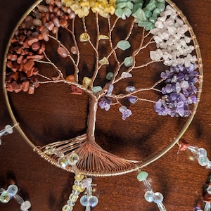 May include: A copper wire tree of life with crystals in the colors of the rainbow. The crystals are attached to the branches of the tree, which is suspended from a hoop. The tree is decorated with crystal beads and hangs from a chain.